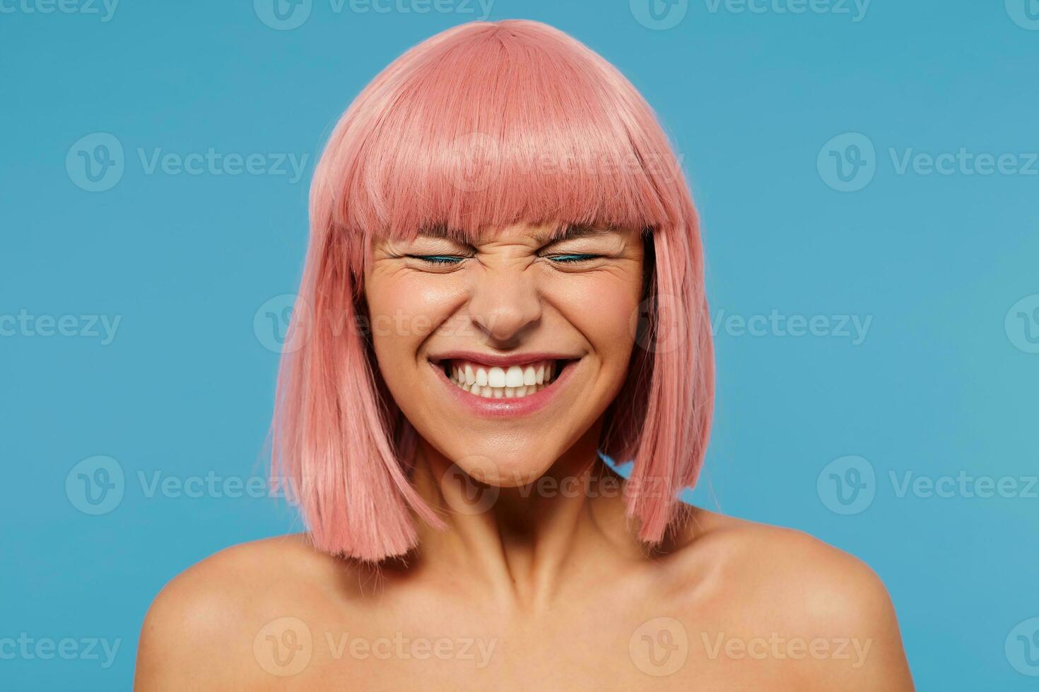 Goog looking young cheerful lovely pink haired female with bob haircut frowning her face while smiling widely, keeping eyes closed while posing over blue background photo