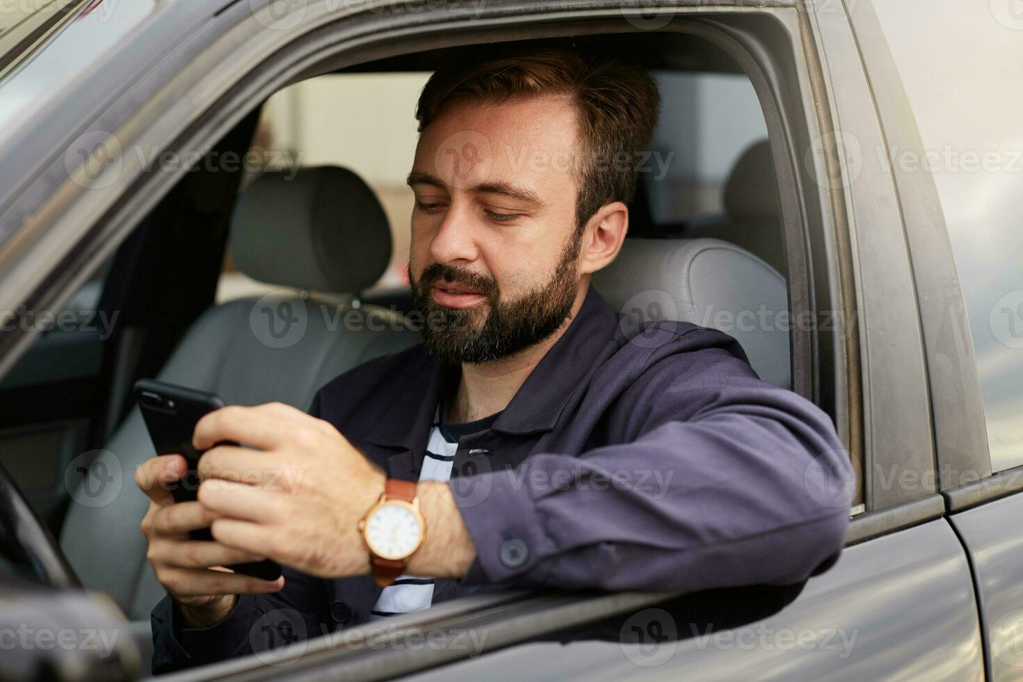 Young hendsome successful bearded man in a blue jacket and striped t-shirt, sits behind the wheel of the car, chatting with colleague by phone. photo