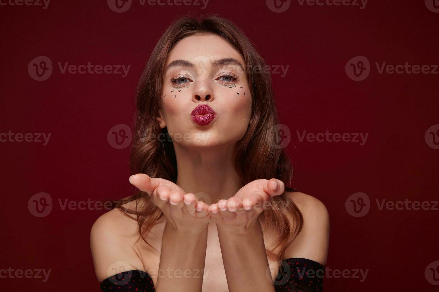 Close-up of cheerful pretty young brown haired woman with festive makeup keeping palms raised while folding lips for air kiss, looking positively at camera while posing over claret background photo