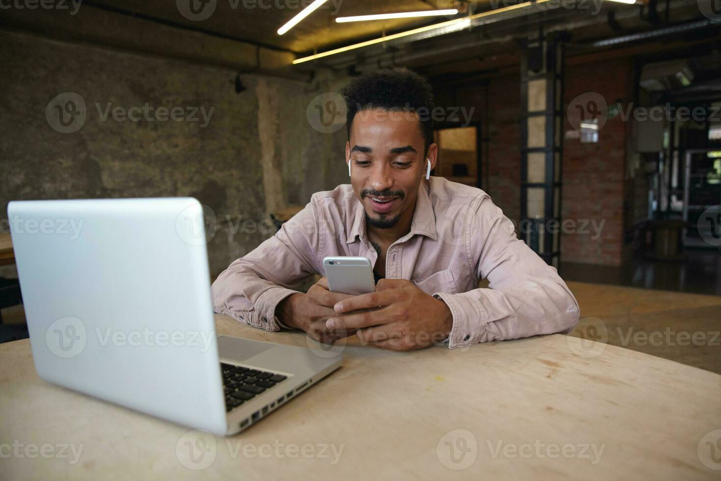 Positive handsome bearded man with dark skin wearing beige shirt, working remotely from coworking space, keeping mobile phone in hands and checking social networks photo
