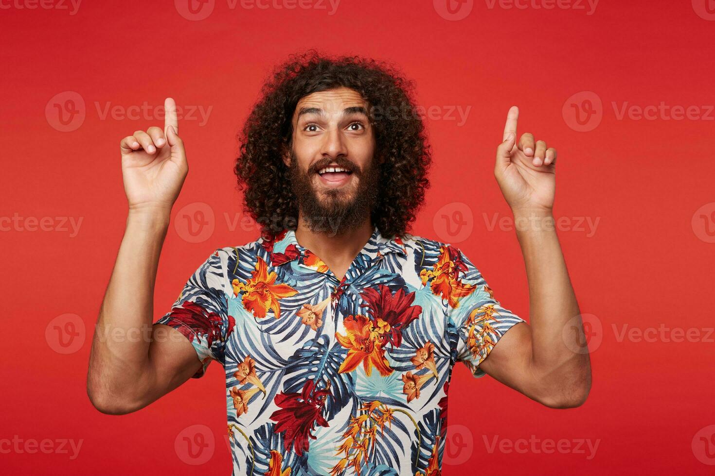 Studio shot of positive young brunette curly man with beard showing up with index fingers while posing against red background in multi-colored flowered shirt photo