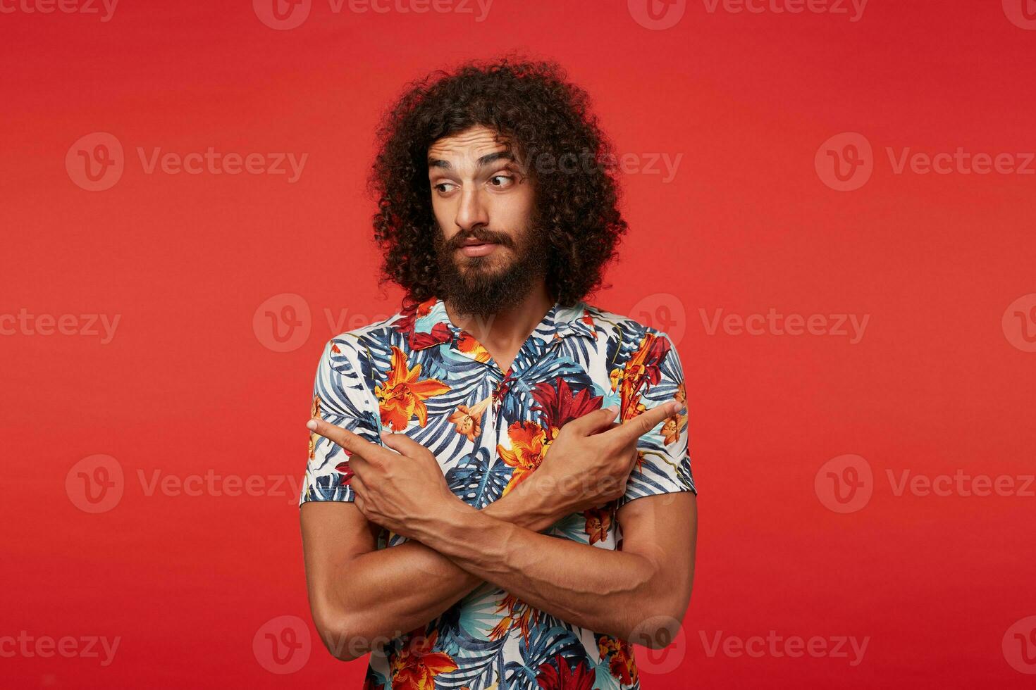 Puzzled young brunette curly bearded man in multi-colored shirt with floral print wrinkling forehead and looking confusedly aside, crossing hands with forefingers showing in different sides photo