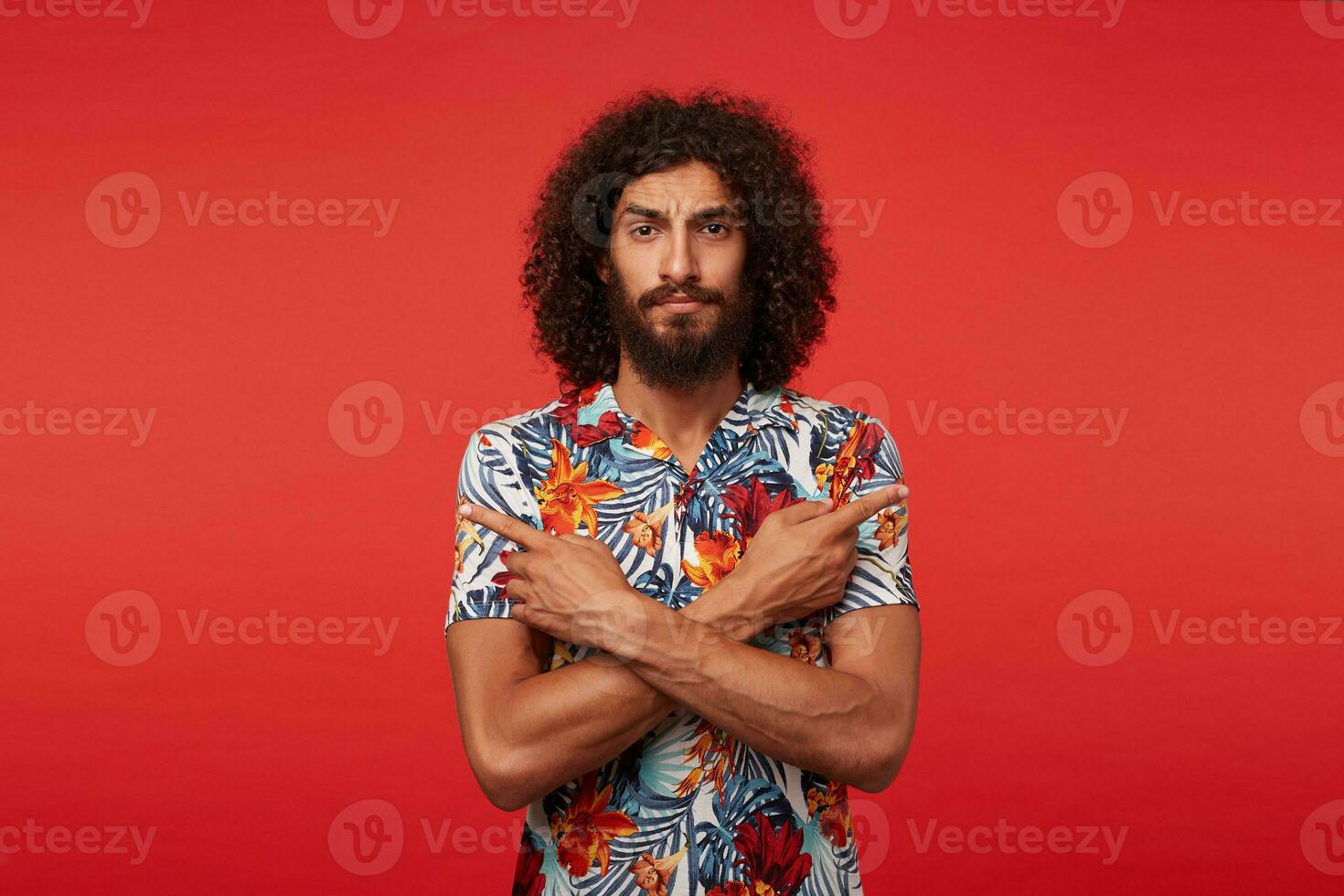 Severe handsome brunette male with lush beard showing in different directions with forefingers, frowning his eyebrows while looking at camera, isolated against red background photo