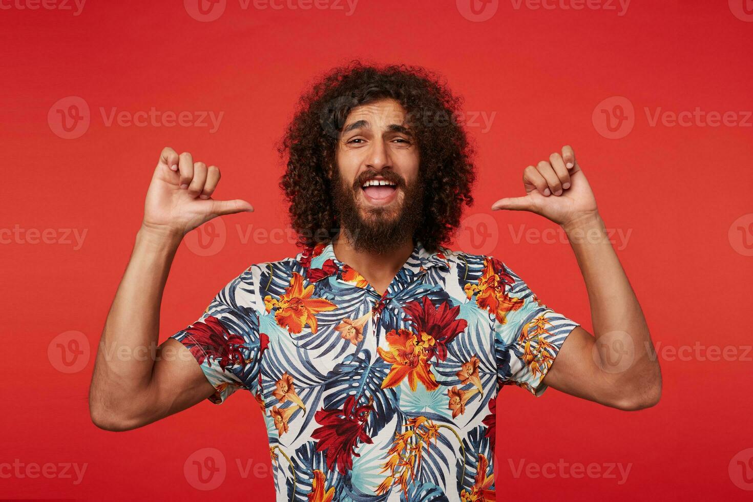 Portrait of young attractive bearded man with brown curly hair looking at camera happily and pointing on himself self-confidently with raised thumbs, isolated over red background photo