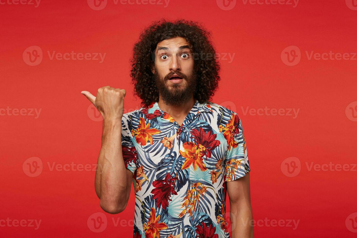 Frightened young pretty bearded man with curly brown hair rounding eyes amazedly and pointing aside with raised hand, wearing multi-colored flowered shirt while posing against red background photo