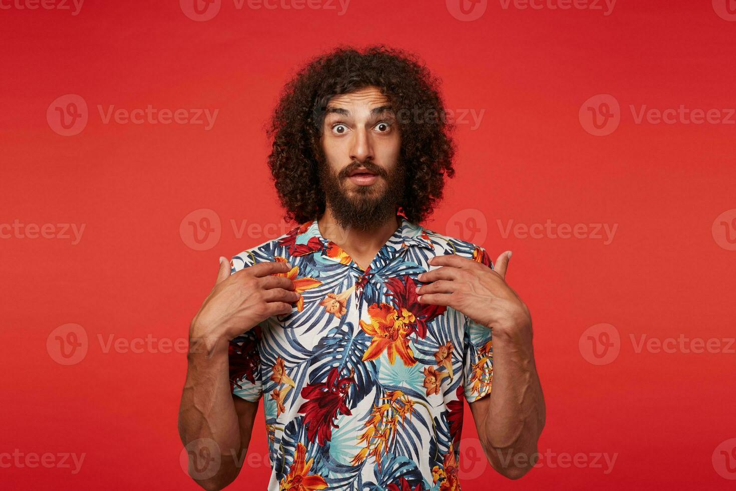 Amazed young brunette curly bearded man showing on himself with raised palms and looking to camera with wide eyes opened, dressed in multi-colored flowered shirt while standing over red background photo