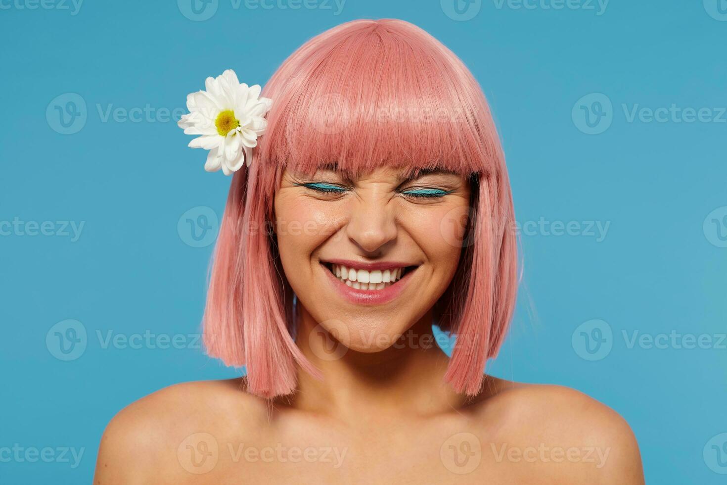 Close-up of cheerful young pink haired lovely female with festive makeup wearing flower in her head while posing over blue background, frowning her face while smiling happily photo