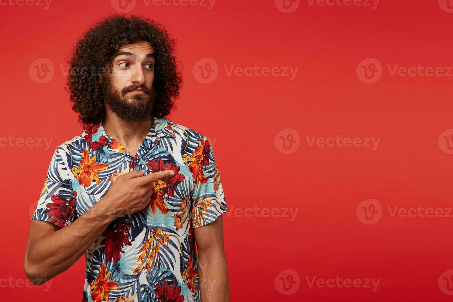 Studio shot of attractive young brunette curly male with beard wearing casual clothes while standing over red background, showing aside with raised forefinger and wrinkling forehead with folded lips photo