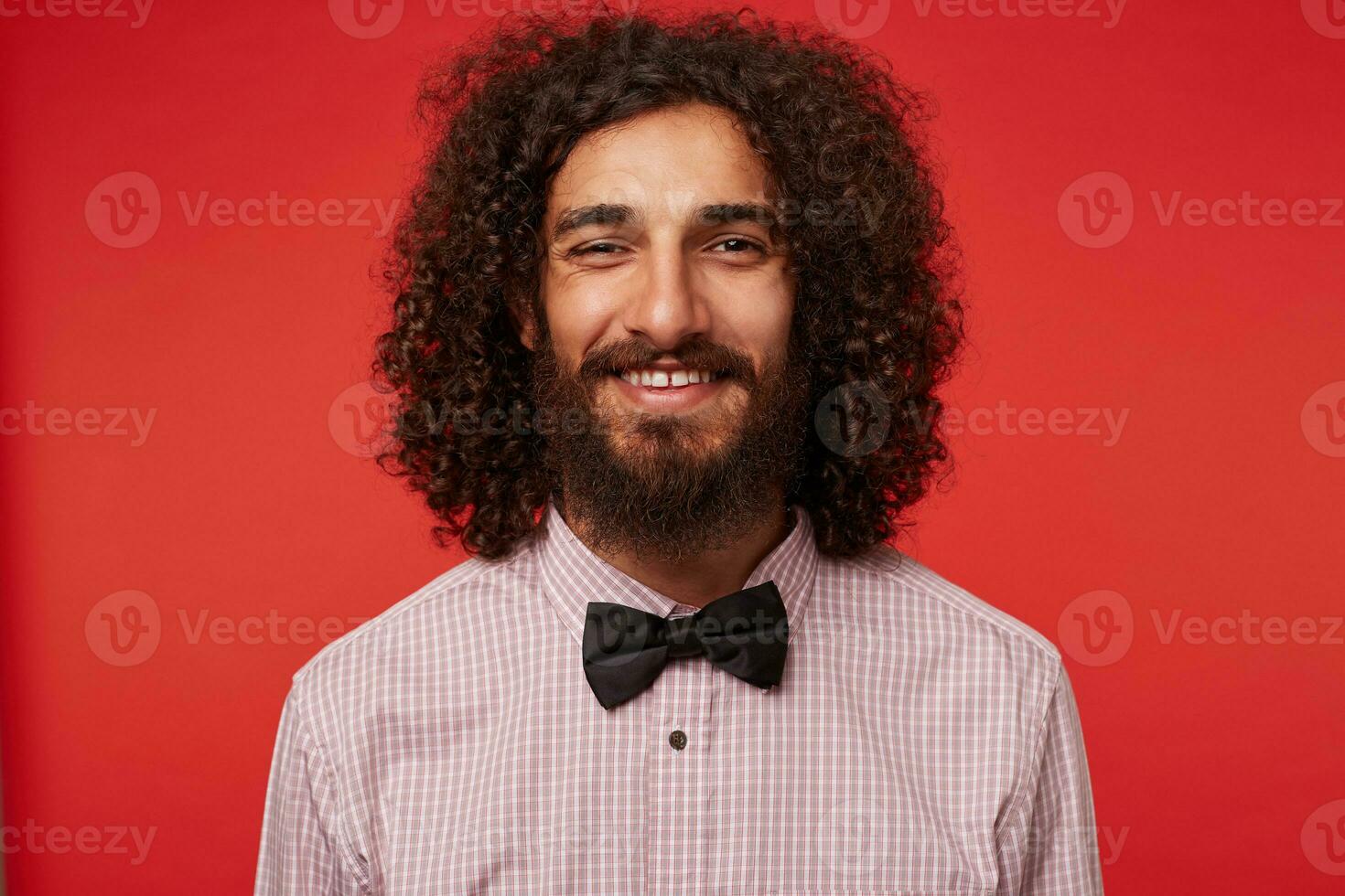 de cerca de encantador joven oscuro peludo Rizado barbado hombre en elegante ropa mirando a cámara con encantador sonrisa y demostración su blanco Perfecto dientes, aislado en contra rojo antecedentes foto