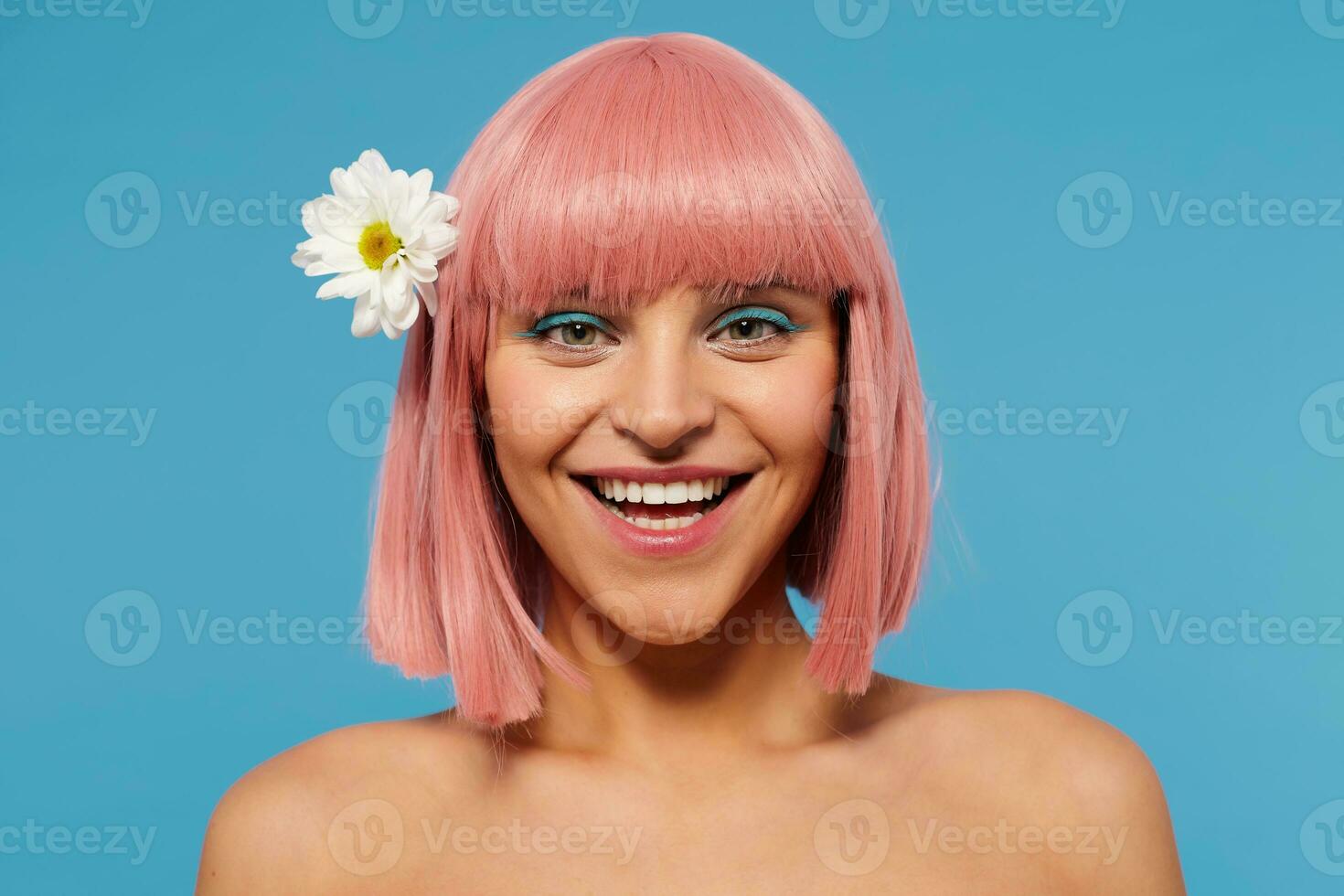 Charming romantic young pink haired lady with colored makeup looking happily at camera and smiling widely, posing over blue background with chamomile in her head photo