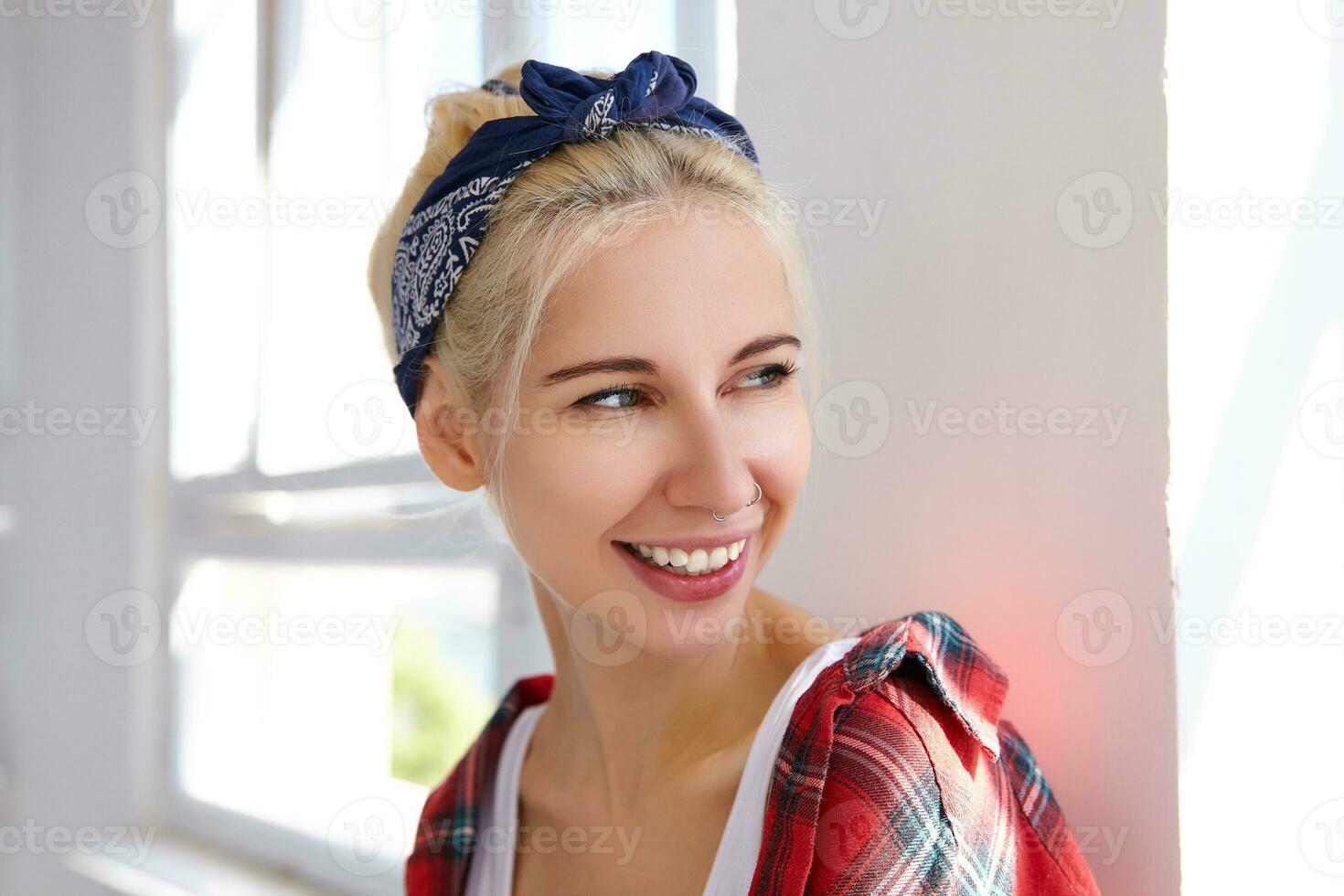 retrato de joven alegre rubia hembra con casual peinado vistiendo venda y casual vestir mientras posando terminado brillante estudio, sonriente extensamente mientras mirando con alegría aparte foto