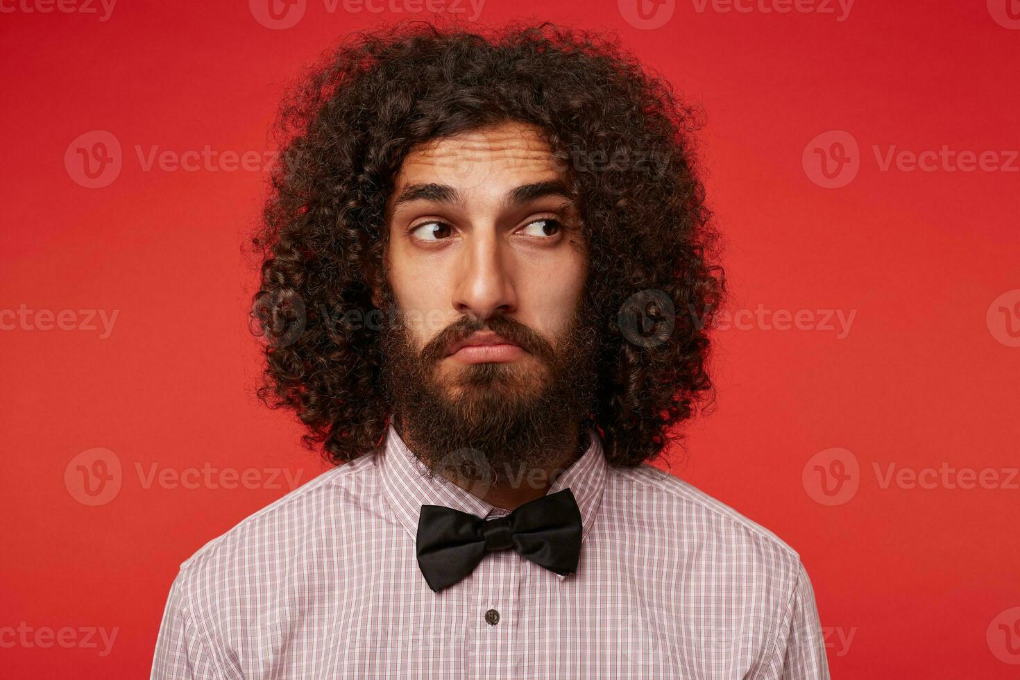 confuso joven morena Rizado masculino con barba arruga frente mientras mirando aparte y retortijón su boca, en pie terminado rojo antecedentes en elegante ropa foto