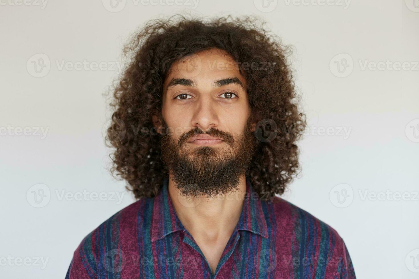 Portrait of young handsome brown-eyed bearded male with dark curly hair looking at camera with calm face, keeping lips folded while standing against white background photo