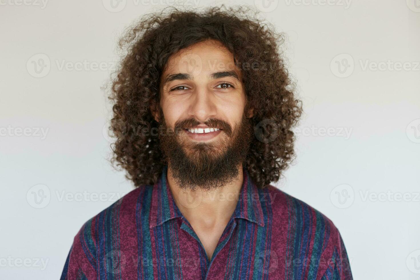 Indoor photo of attractive young dark haired bearded man looking positively at camera with charming smile, showing his white perfect teeth while posing against white background