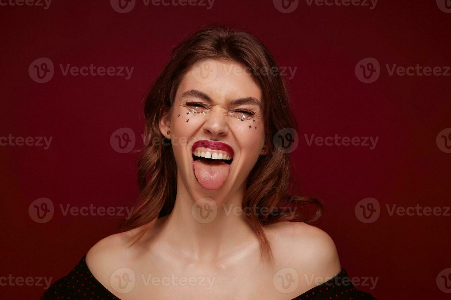 Close-up of joyful excited young brunette woman with silver stars on her face showing tongue to camera and squinting her eyes, posing over claret background in top with opened shoulders photo