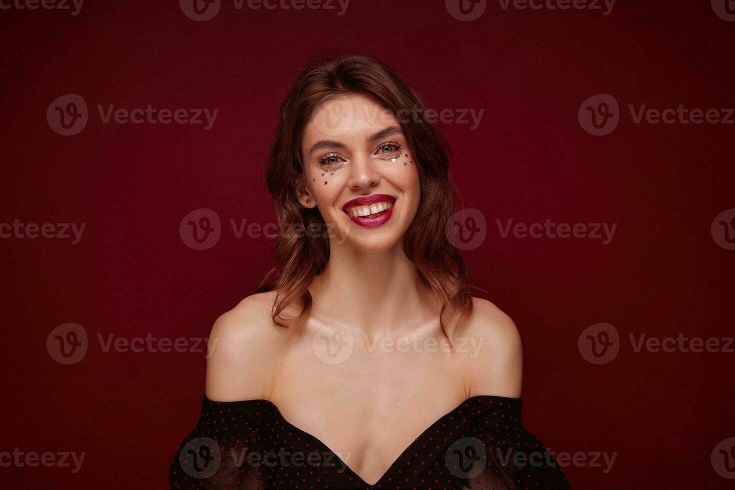 Indoor shot of attractive young brown haired female with wavy hairstyle looking cheerfully at camera and demonstrating her white perfect teeth, isolated over claret background photo