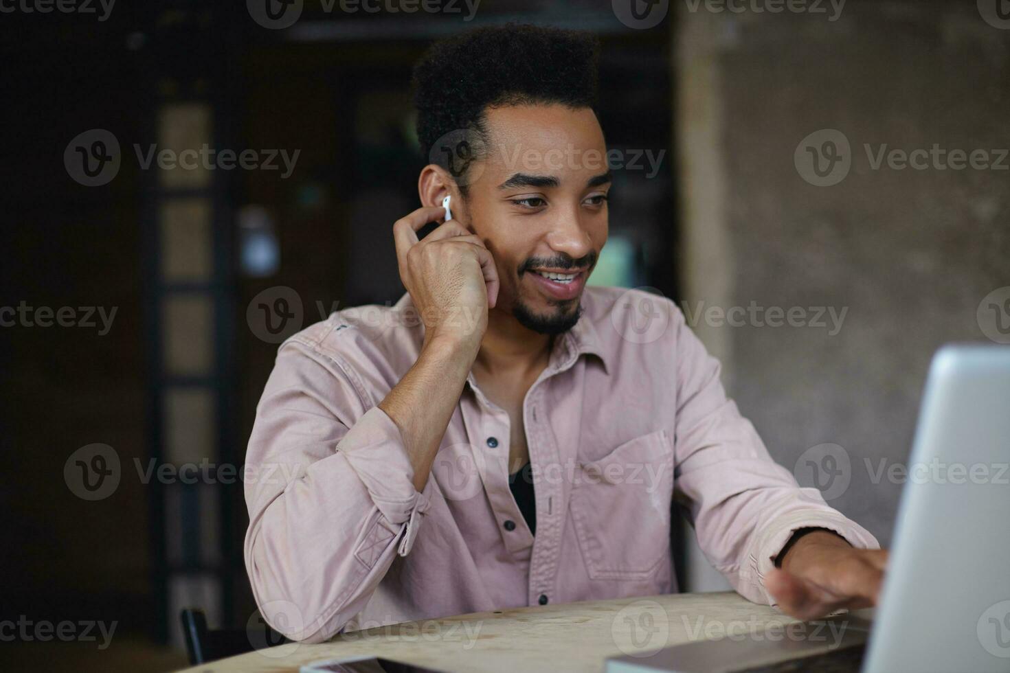Positive smiling attarctive young bearded guy with dark skin sitting over city cafe and watching funny videos with earphones while having lunch, wearing casual clothes photo