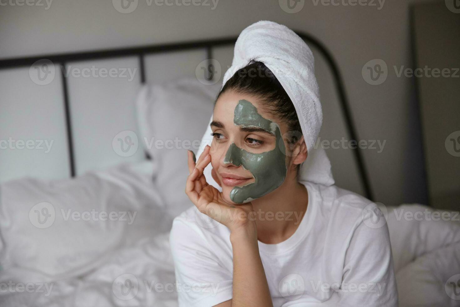 Portrait of attractive young dark haired woman with towel on her head leaning chin on raised hand and looking aside thoughtfully while keeping mask on her face, sitting bedroom interior photo