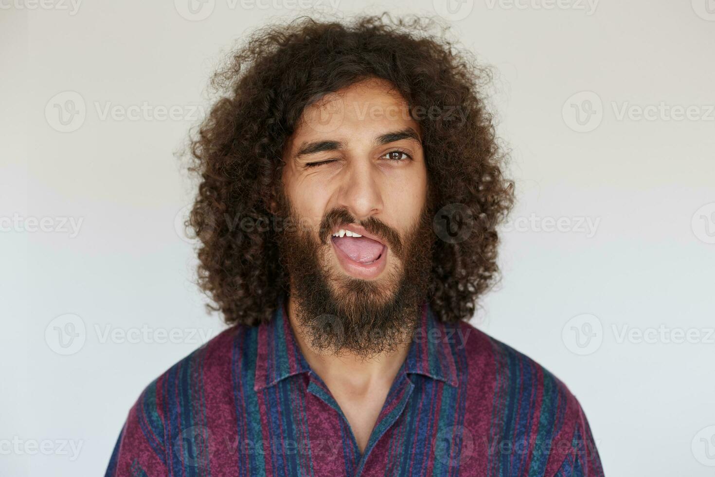Indoor shot of young brunette curly male with lush beard giving wink to camera with wide mouth opened, wearing striped multi-colored shirt against white background photo