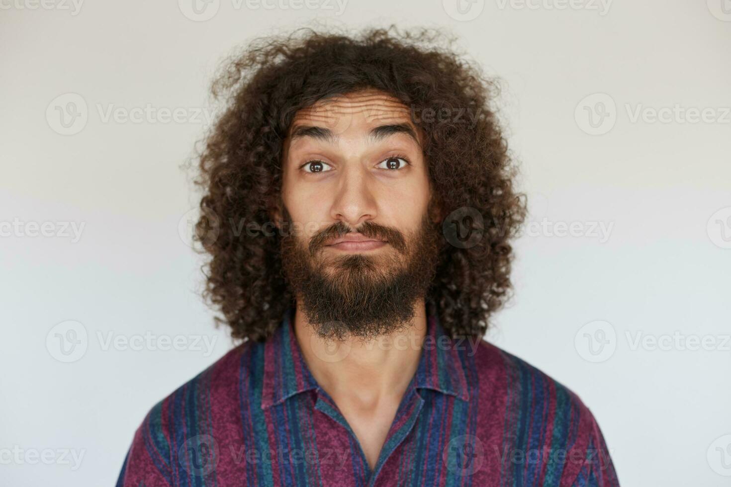 Studio photo of attractive brown-eyed young bearded male with dark curly hair raising eyebrows surprisedly while looking at camera, dressed in striped multi-colored shirt against white background