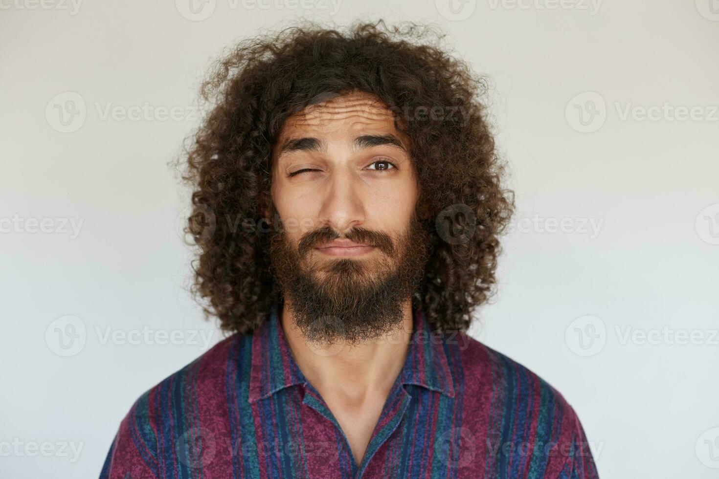 Portrait of pretty young dark haired curly bearded man with folded lips keeping eye closed while looking to camera, wearing casual clothes while standing over white background photo