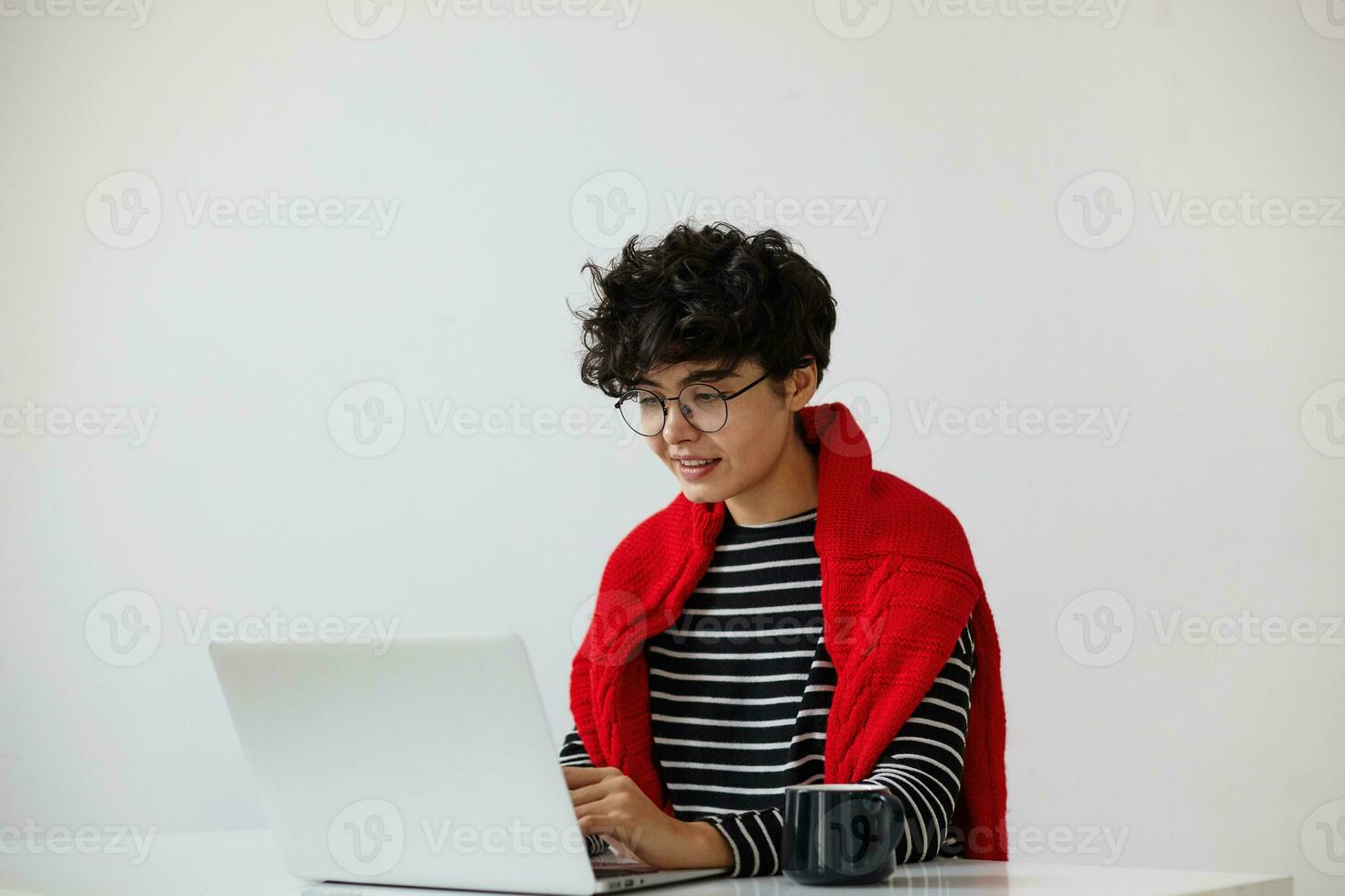 positivo joven atractivo mujer con corto Rizado pelo vistiendo los anteojos mientras trabajando a moderno oficina con computadora portátil, mirando a pantalla mientras mecanografía carta, aislado terminado blanco antecedentes foto