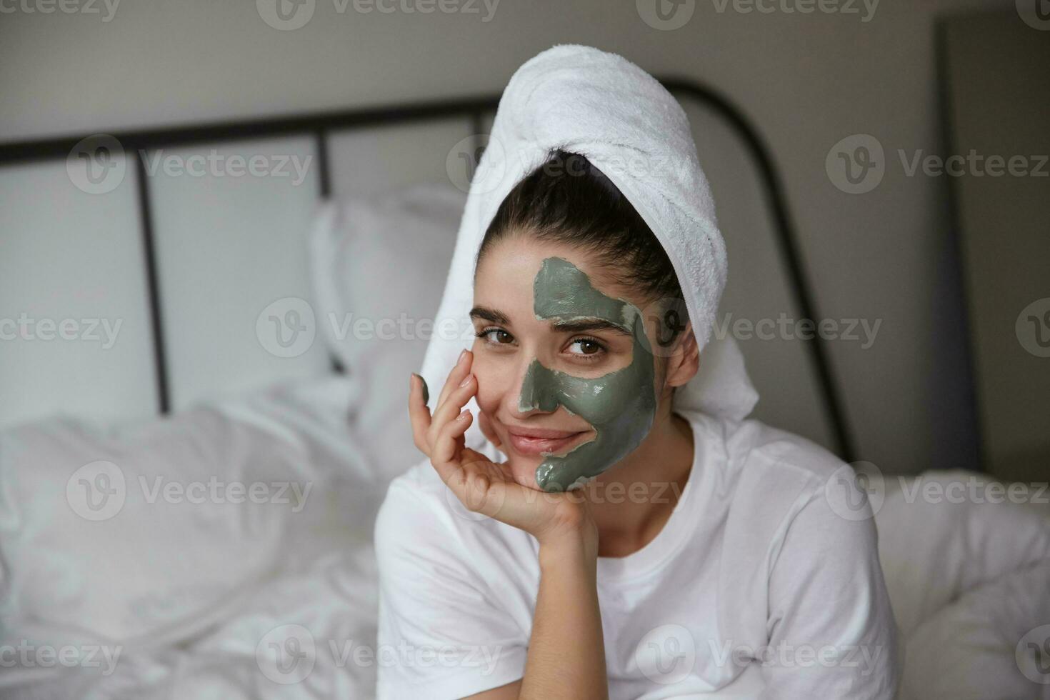 Cosy photo of young pretty dark haired woman with brown eyes holding her chin on raised hand and looking at camera with light smile, making skin care procedure at home