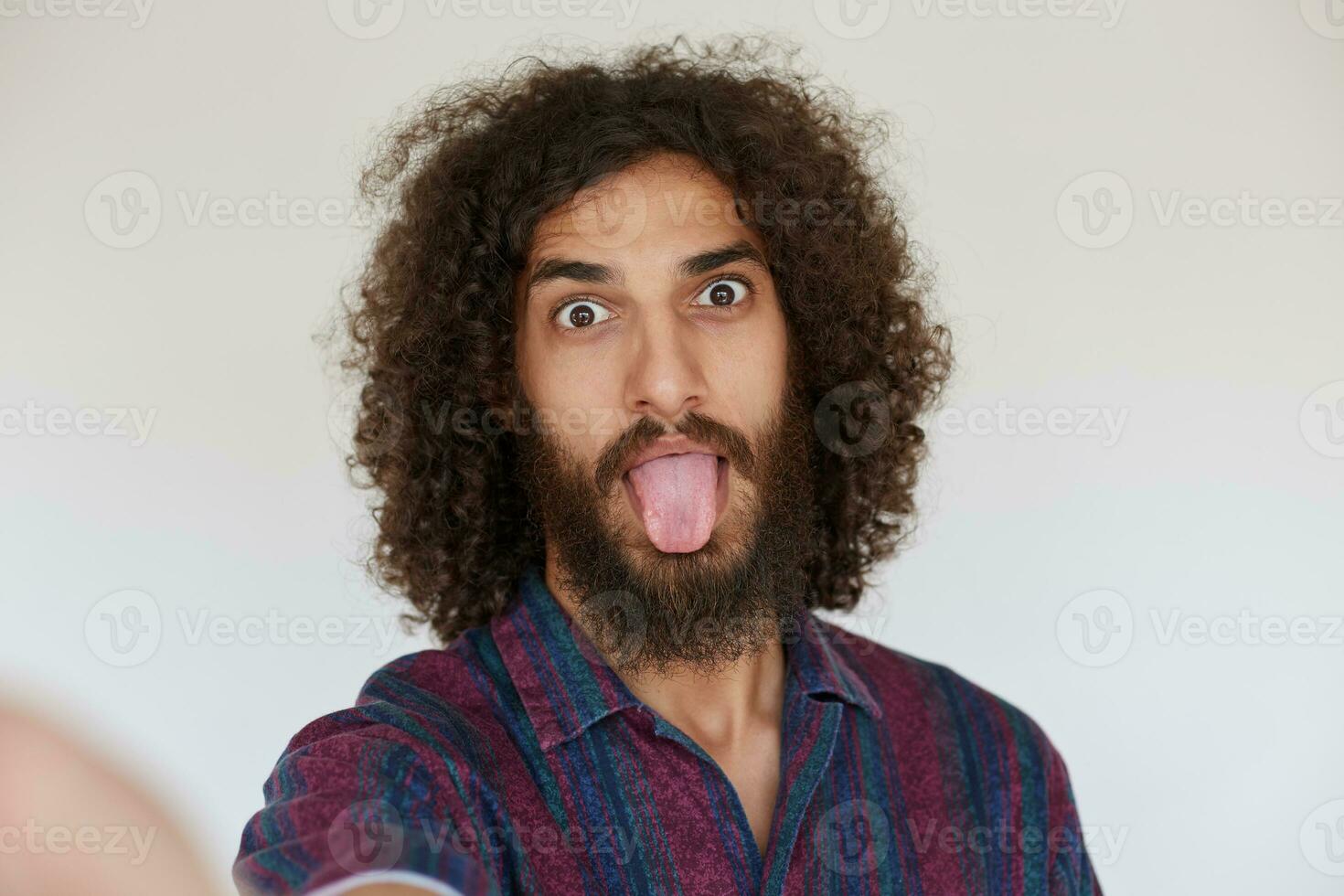Funny indoor shot of attractive young dark haired curly bearded man fooling and making faces while standing against white background, dressed in striped multi-colored shirt photo