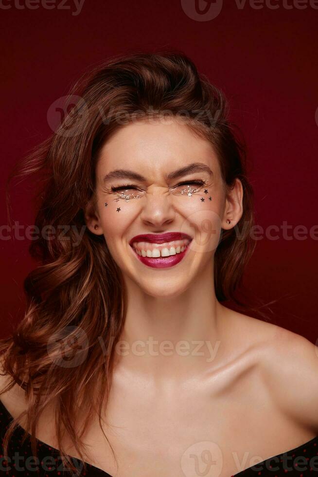 Beautiful cheerful young lady with brown wavy hair laughing happily and showing her white perfect teeth, wearing evening makeup and small silver stars on her face, isolated over claret background photo