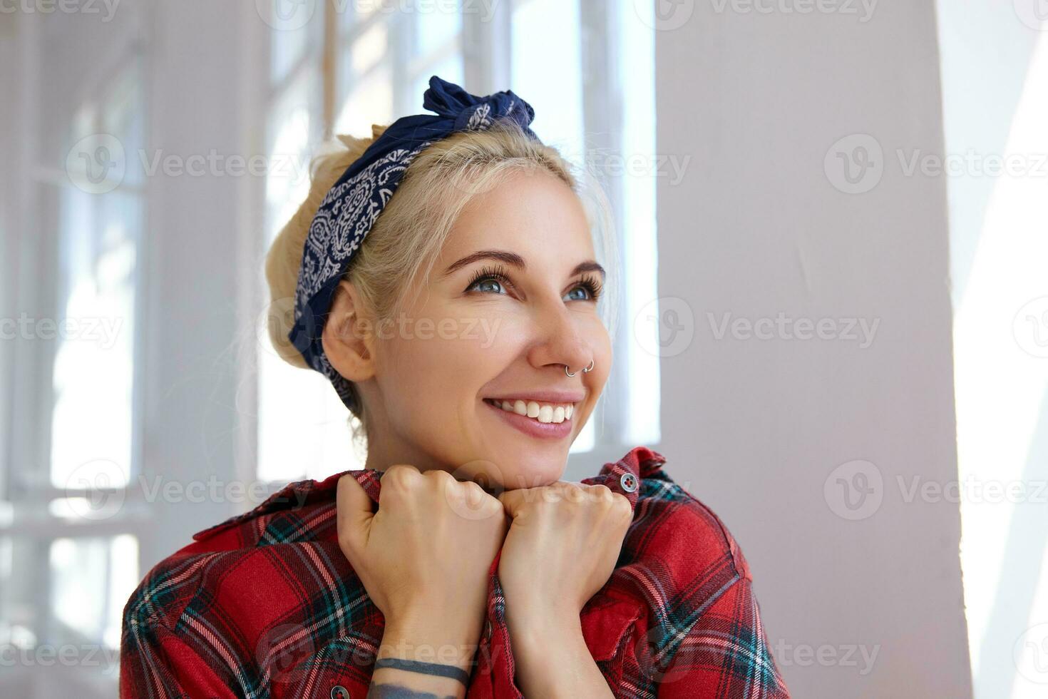 Close-up of young happy blonde in headband leaning her face on raised hands and smiling cheerfully, being in nice mood while standing in front of big window photo