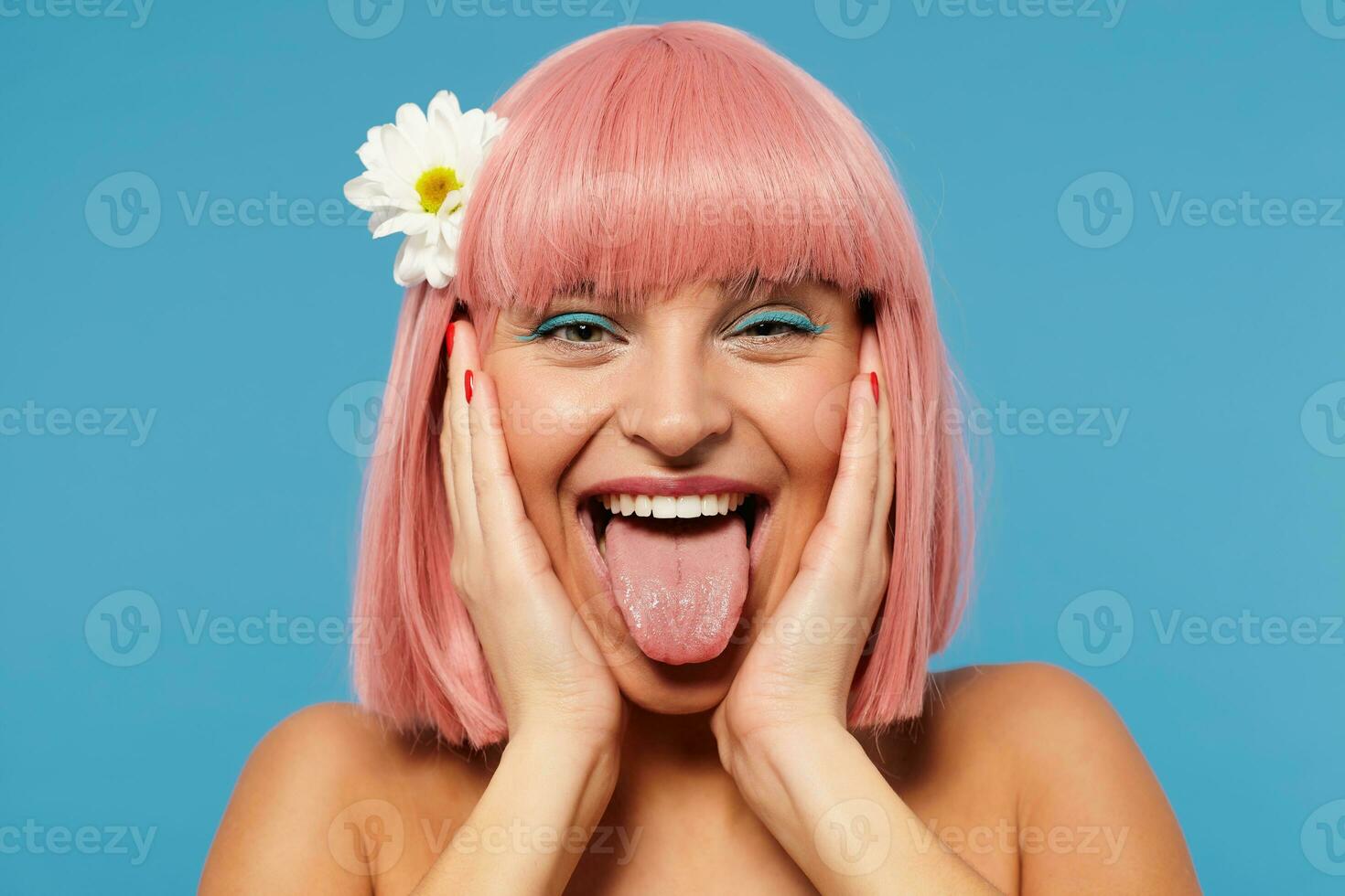 Funny shot of cheerful young lovely pink haired woman with trendy haircut holding her face with raised palms while posing over blue background, fooling and showing tongue at camera photo