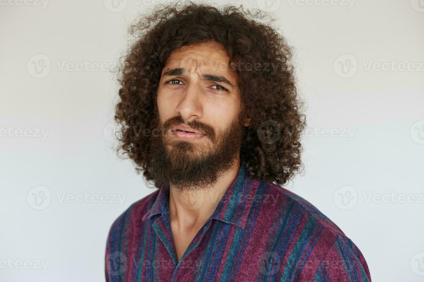 Discontented young dark haired curly male with lush beard frowning eyebrows with pout while standing against white background in striped multi-colored shirt photo