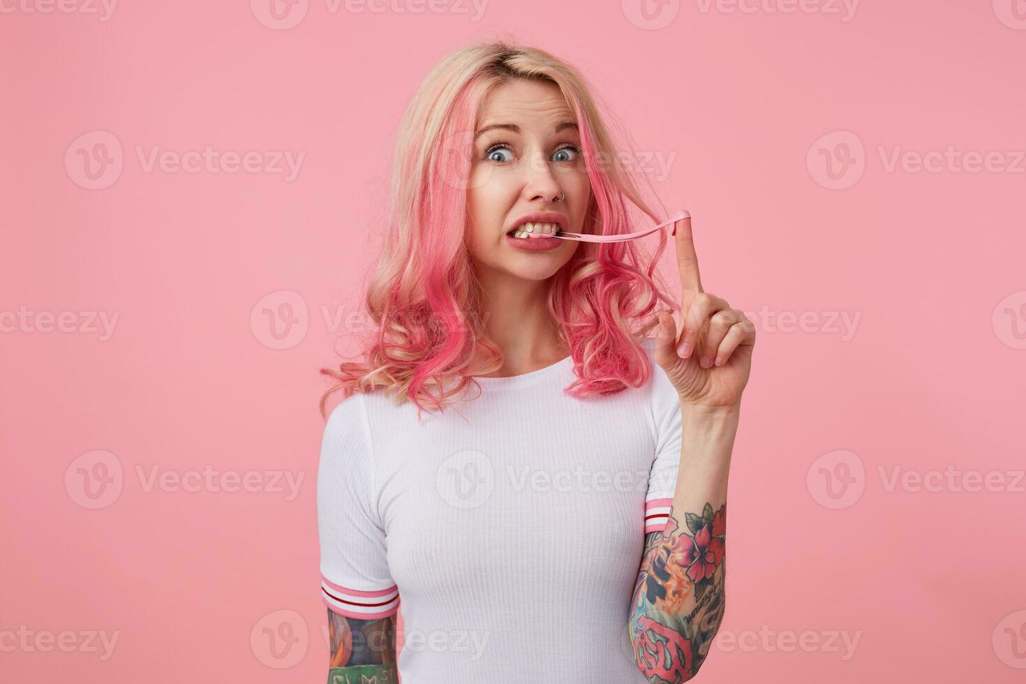 retrato de joven preguntado linda rosado peludo dama con tatuado manos, usa en blanco camiseta, disfruta fresa burbuja chicle, confuso mira a el cámara soportes terminado rosado antecedentes. foto