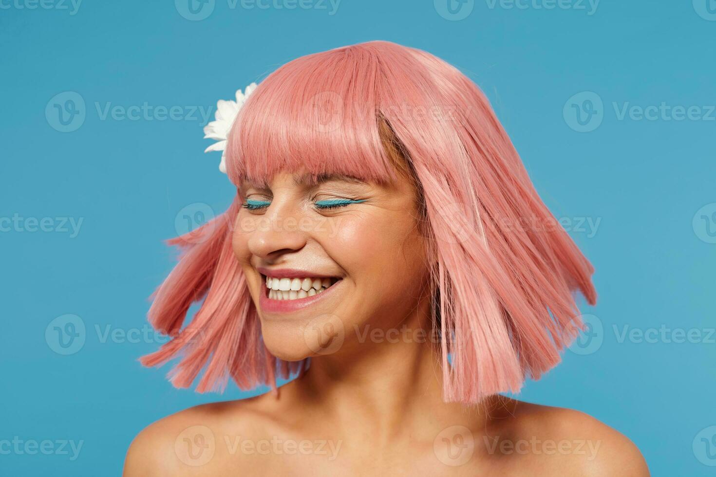 Good looking happy young romantic lady with short pink haircut waving her hair and smiling cheerfully with closed eyes, standing against blue background with naked shoulders photo