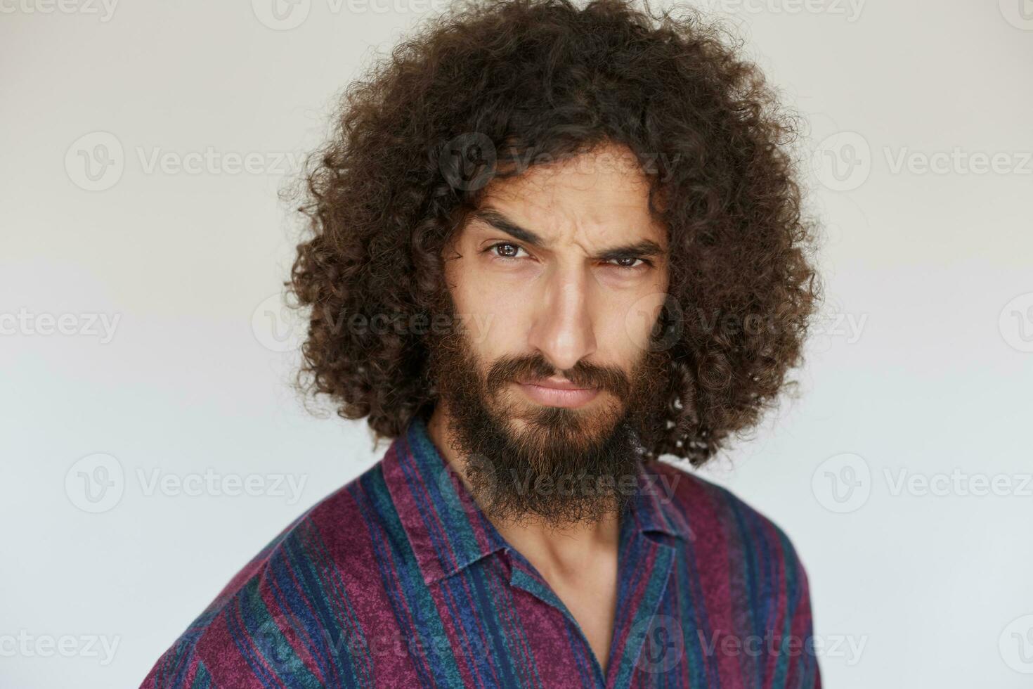 Portrait of stern brunette bearded guy with dark curly hair raising eyebrow and looking severely at camera, keeping lips folded while posing against white background in casual shirt photo