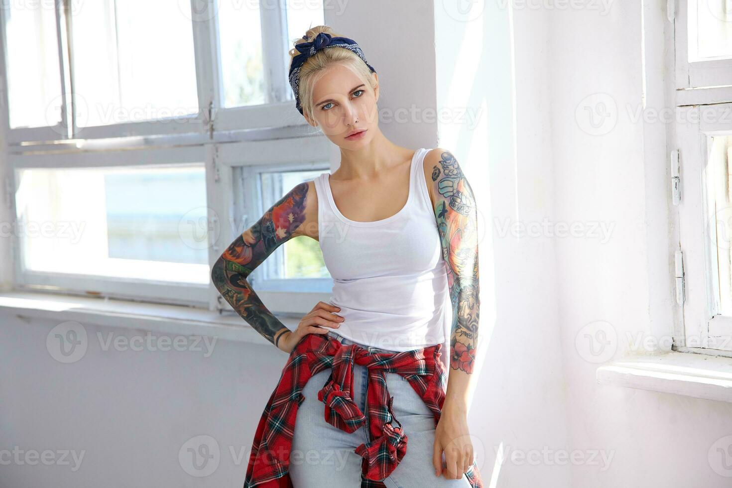 Severe young attractive tattooed blonde female with natural makeup keeping her lips folded while looking seriously at camera, posing over bright studio photo