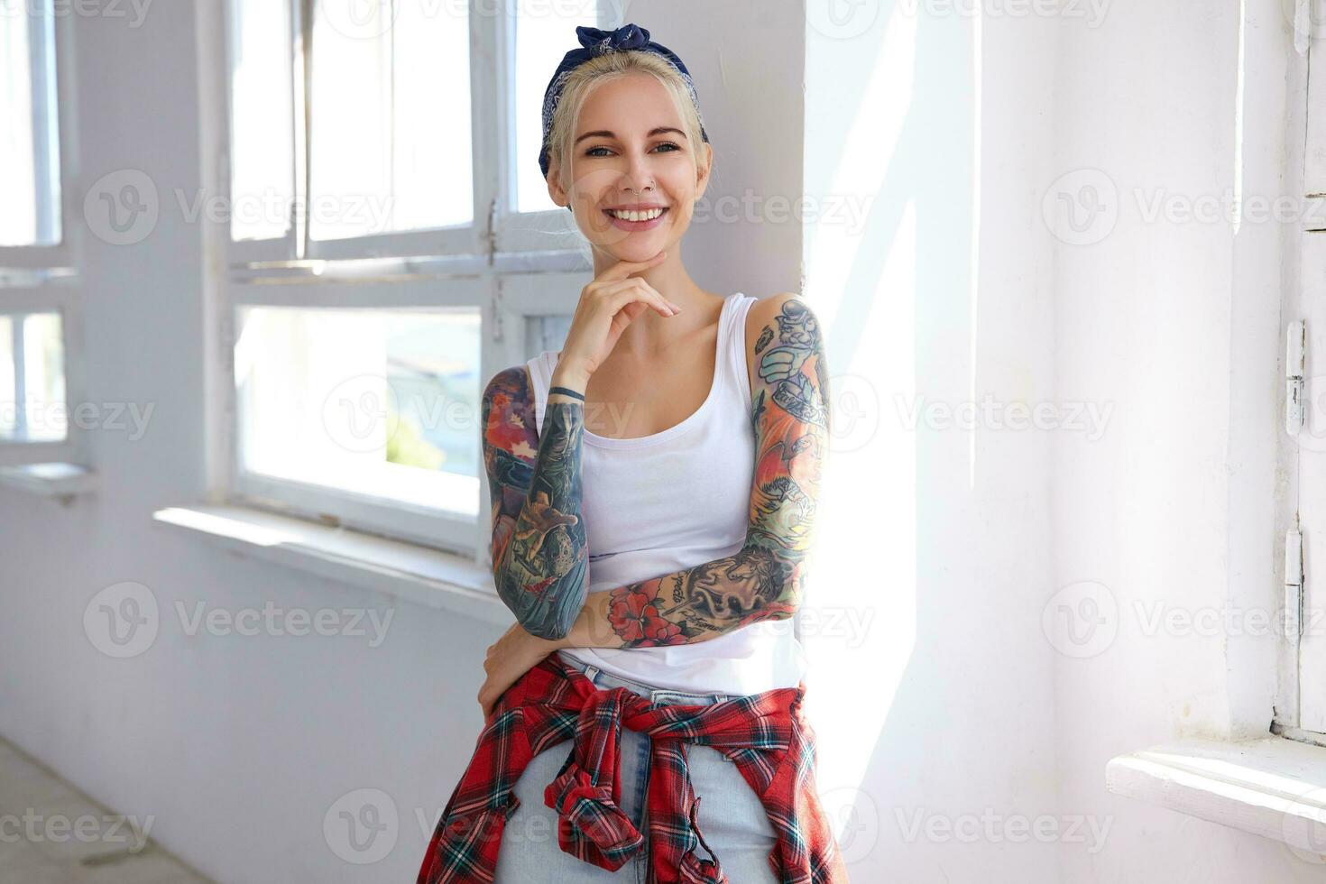 Cheerful young tattooed attractive female in headband and casual wear touching her chin with raised hand while looking gladly at camera with pleasant smile photo