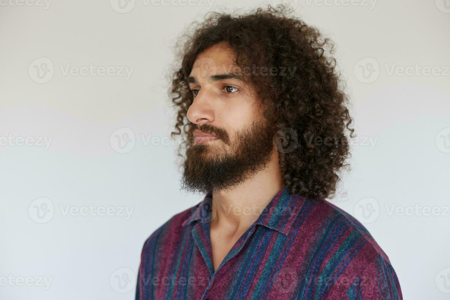 Close-up of attractive bearded guy with dark curly hair looking aside with calm face and keeping his lips folded, dressed in casual shirt while standing against white background photo