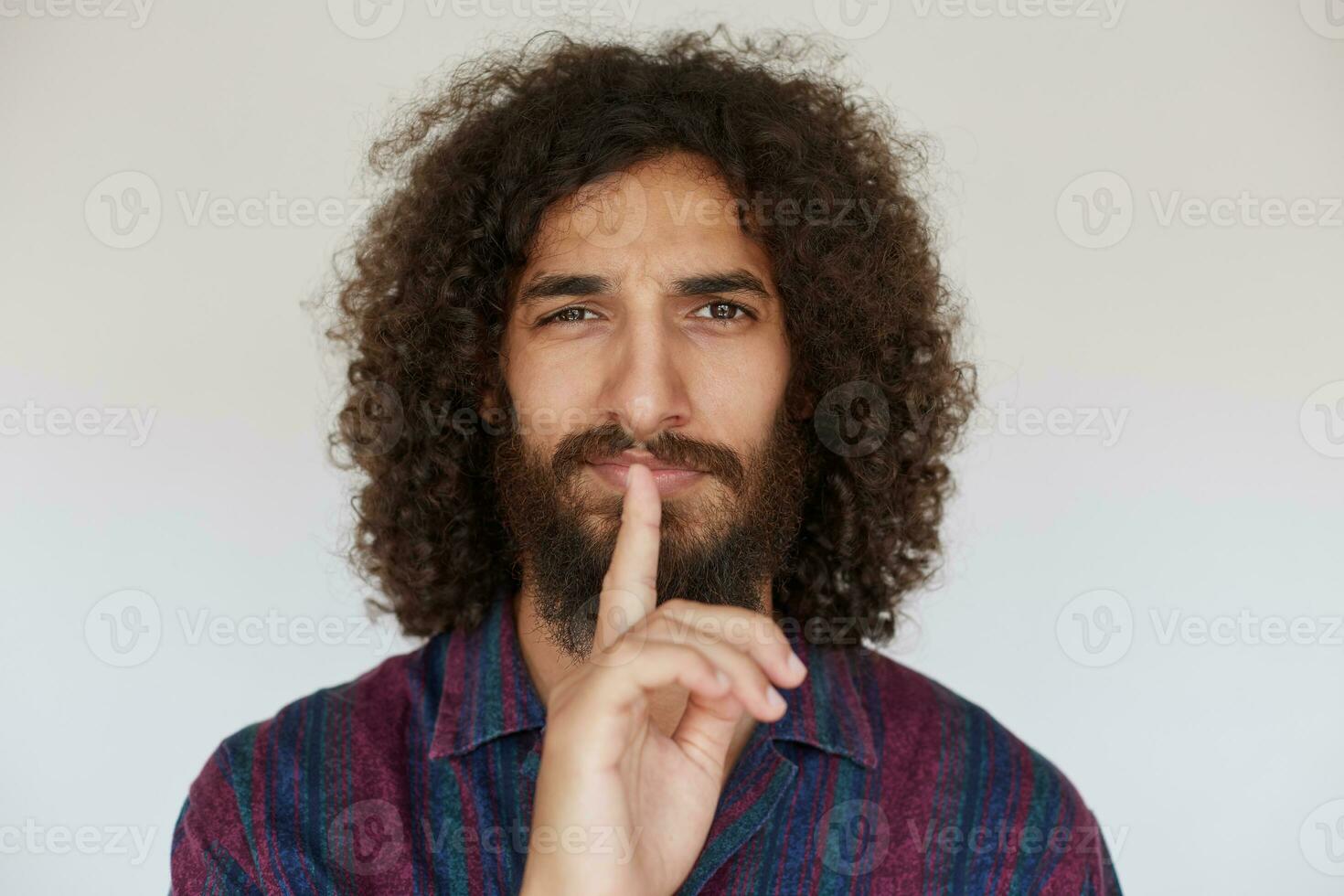 Indoor photo of lovely brunette bearded man in striped multi-colored shirt raising hand in hush gesture, keeping forefinger on his lips while posing over white background