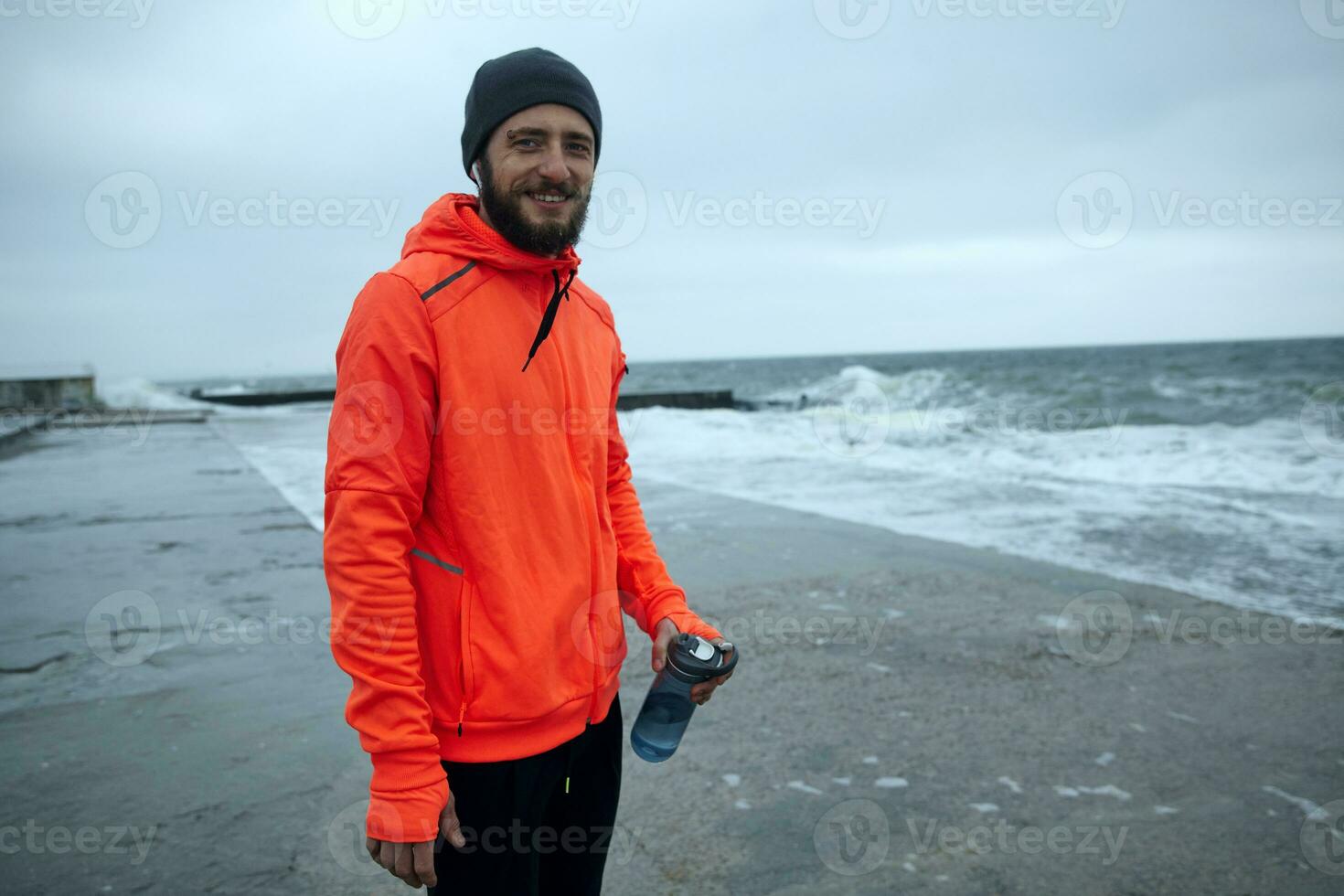 Shot of young cheerful bearded sporty male with eyebrow piercing holding bottle with water in his hand while standing over coastline of sea on stormy cold weather, smiling positively to camera photo