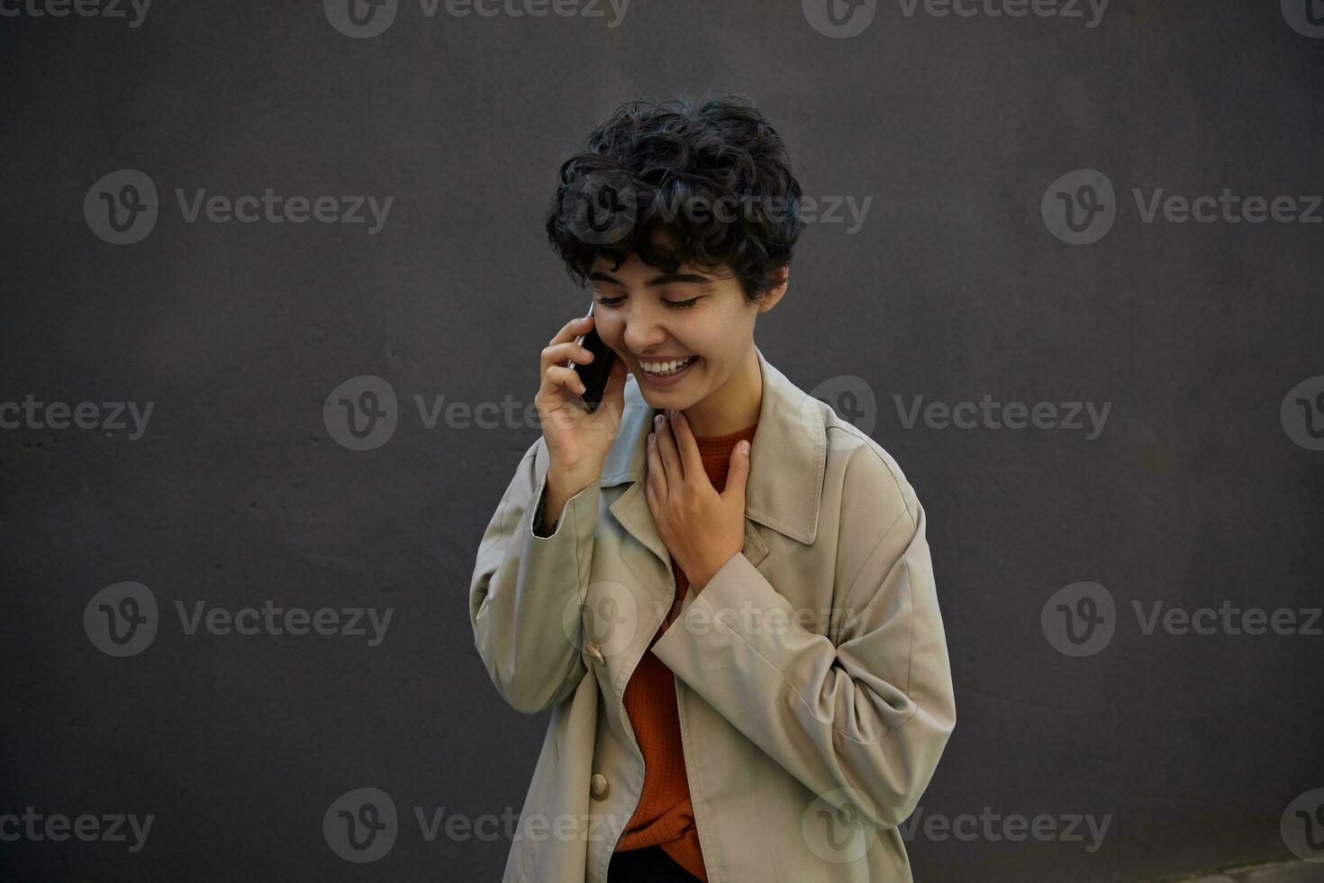 Outdoor photo of pretty young dark haired curly woman with charming smile holding mobile phone and having nice talk, keeping palm on her chest while posing over city environment