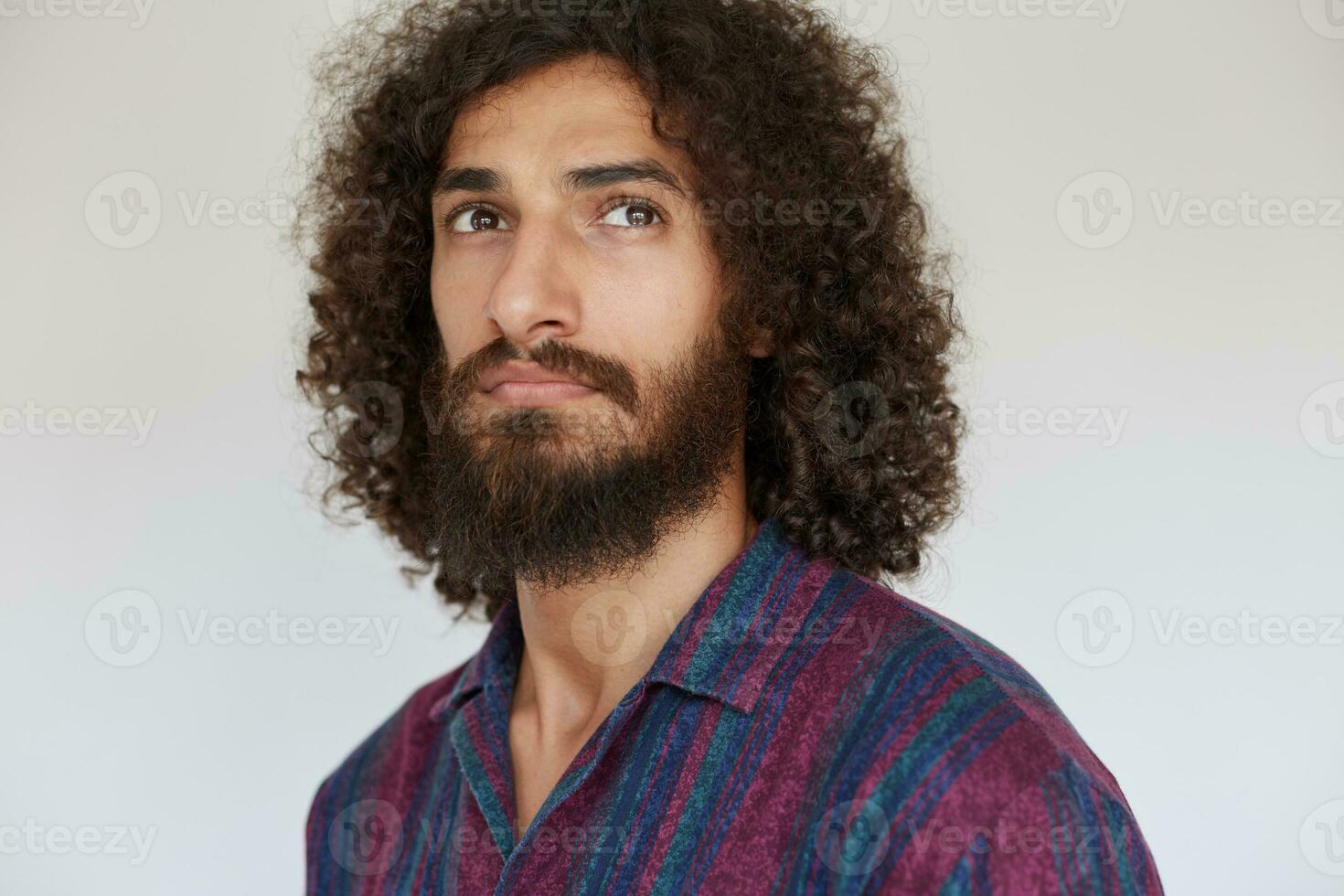 Horizontal shot of good looking serious brunette curly bearded guy with charming brown eyes wearing striped multi-colored shirt while posing against white background photo