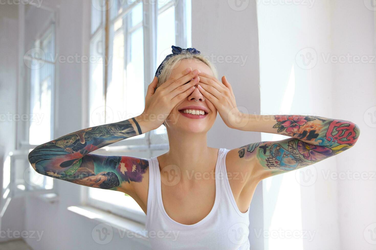 Glad good looking young blonde female with tattoos holding palms on her eyes and smiling widely while expecting surprise, being in high spirit while posing over bright studio photo