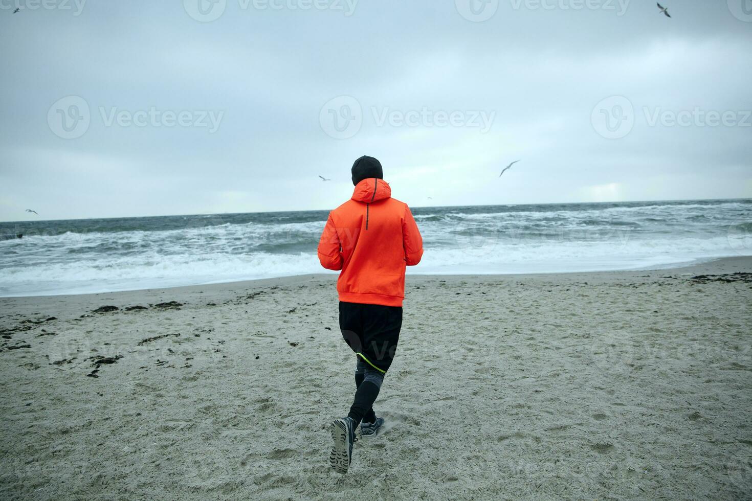 Back view of fitness male model working out and running by seaside on gray early morning, dressed in warm sporty clothes and orange coat. Sport and healthy lifestyle concept photo