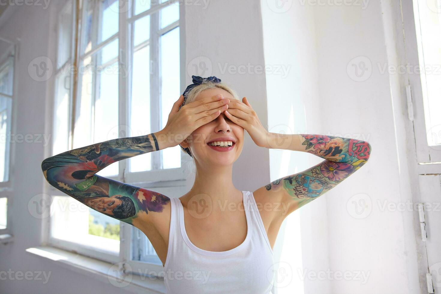 Good looking young lovely glad blonde woman with tattoos keeping raised palms on her eyes and smiling happily while standing over bright studio in white shirt photo