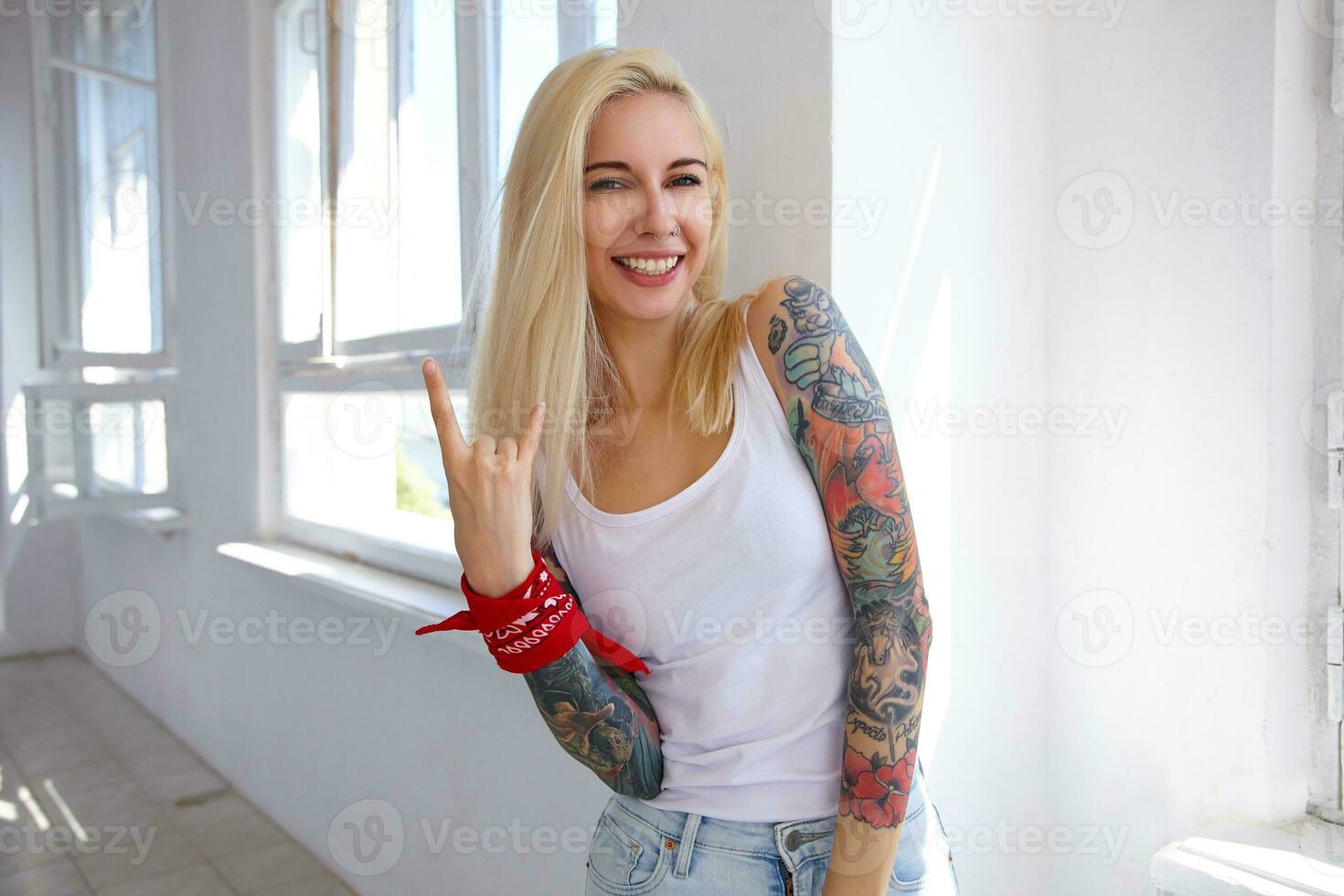 Indoor photo of young blonde long haired female with tattoos making rock and roll sign with raised hand and smiling cheerfully at camera, posing in front of big window