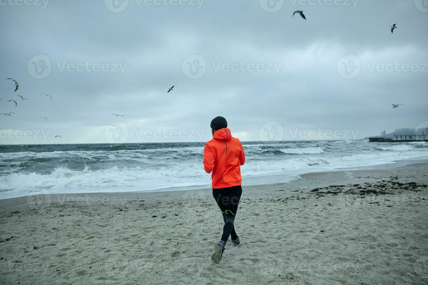 Photo of active sportsman in warm athletic clothes doing physical exercises outdoor, running by seaside before working day, leads healthy sporty lifestyle