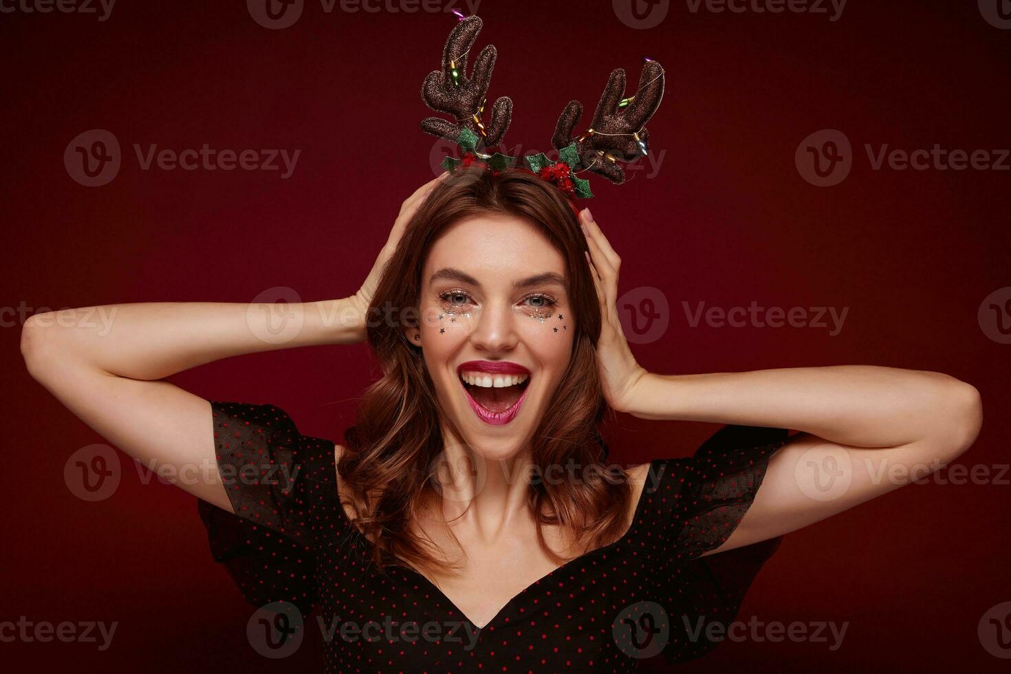 Joyful attractive young brunette woman holding hands on her head and looking happily to camera, preparing for christmas celebration and wearing holday hoop, isolated over claret background photo