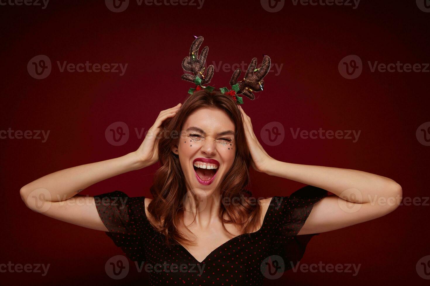 Overjoyed pretty young brunette woman with wavy hairstyle dressed in elegant clothes rejoicing while posing against claret background, wearing funny holiday hoop and laughing happily photo