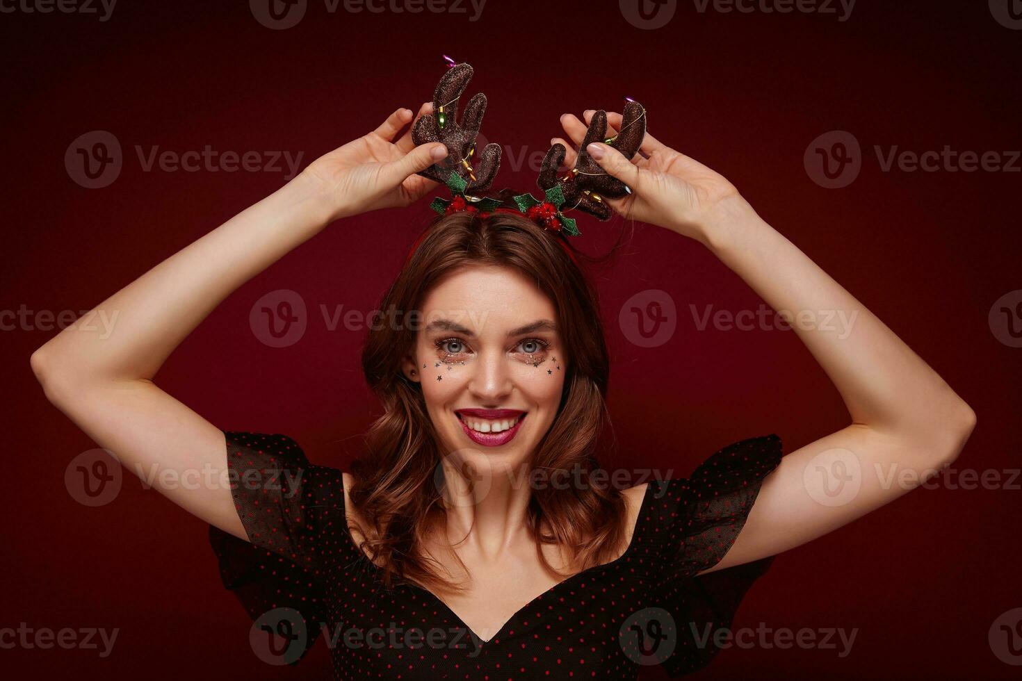 Studio shot of charming young brown haired female in festive clothes raising hands to head horns and smiling widely to camera, enjoying x-mas theme party, isolated over claret background photo